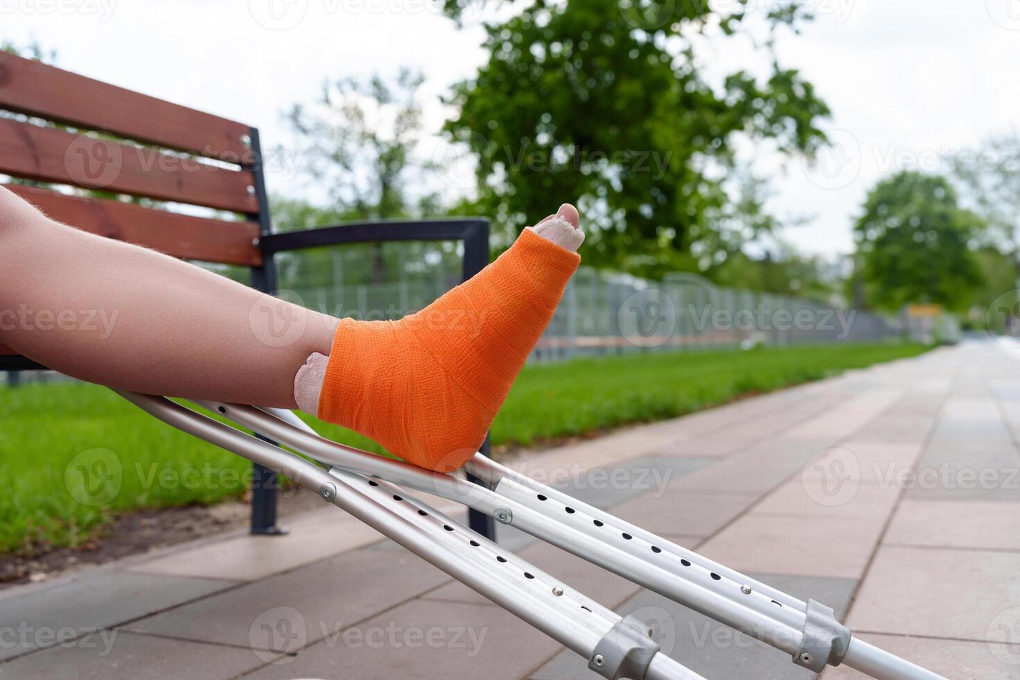 Close-up of cast-encased leg in orange bandage resting on metal crutch, conveying mobility challenges in summer park setting, for healthcare, medical equipment, disability photo