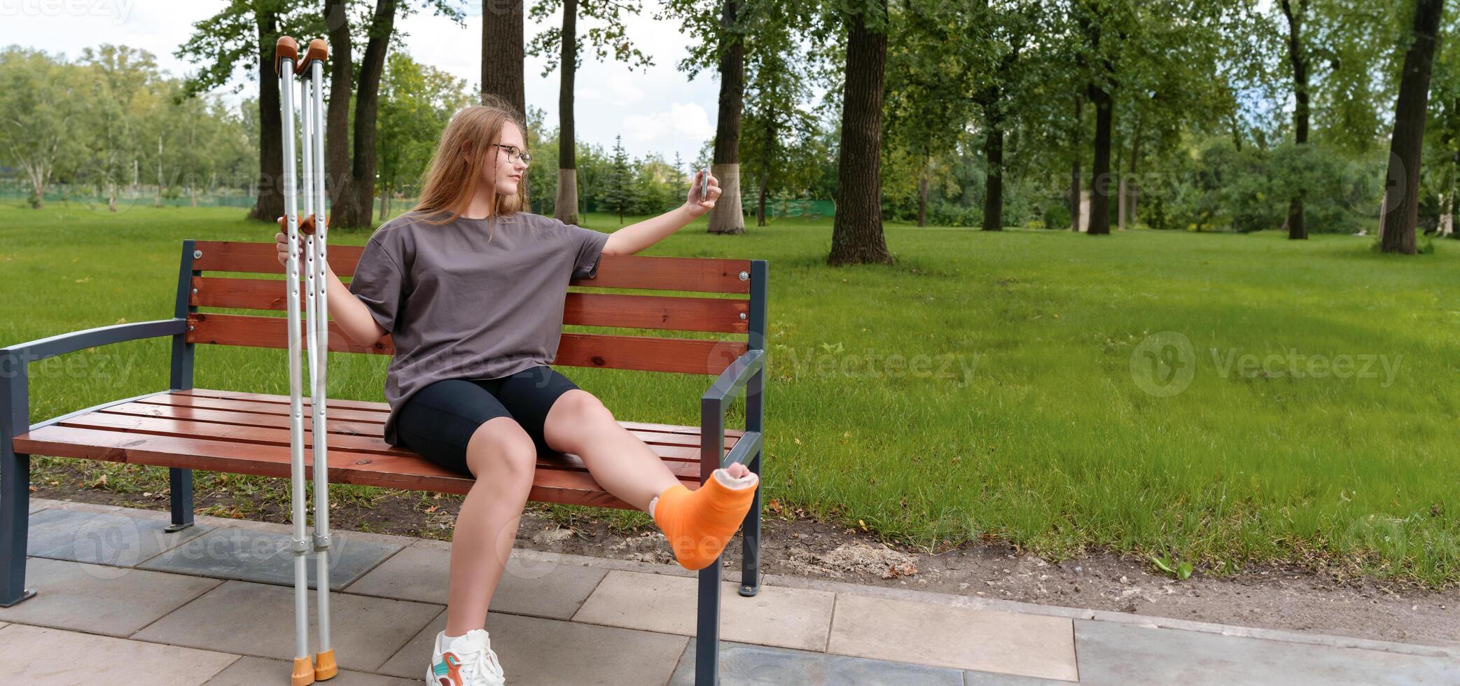 Teenage girl with a cast on her leg seated on a park bench, taking a thoughtful selfie to document the difficulties of her injury and the time she's had to spend in recovery during the summer photo