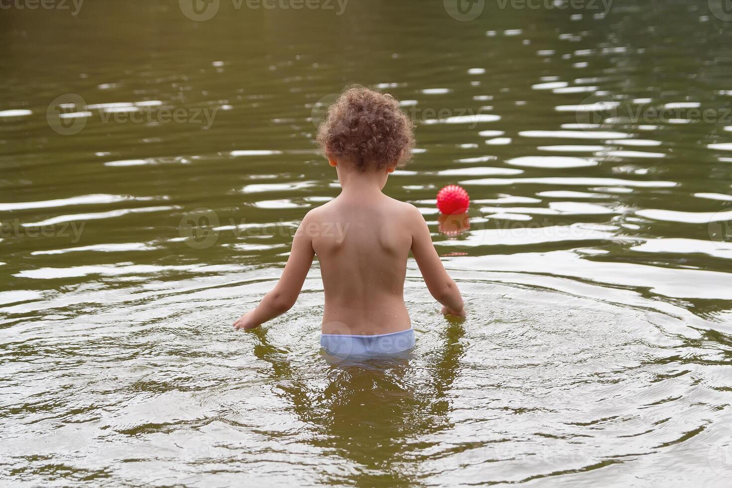 A curly child plays with a ball in the lake in the summer photo