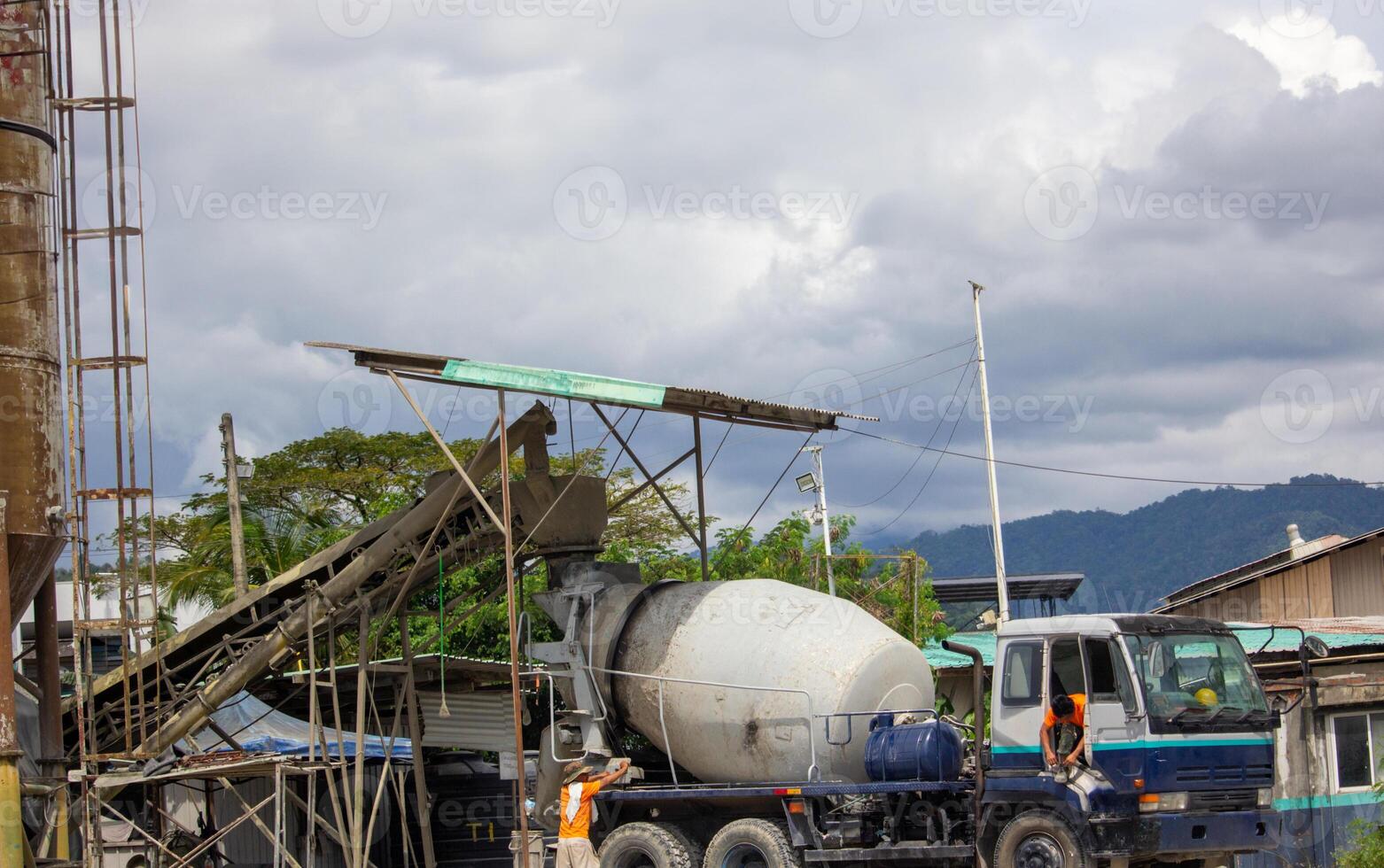 Concrete mixer truck loading at batching plant in industrial site with cloudy sky and mountains photo