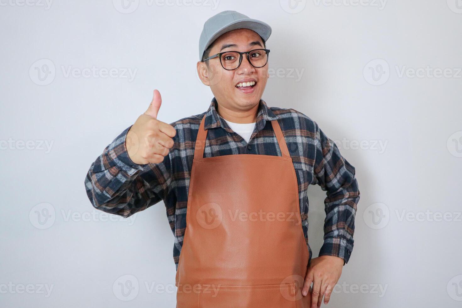 Smiling young Asian man barista employee wearing brown apron working in coffee shop, Looking on camera showing thumbs up in approval and recommend on white background. Small business startup concept photo