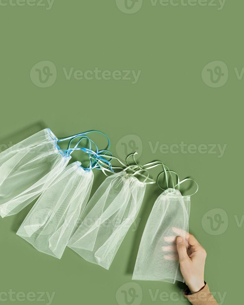 Hand holding reusable grocery bags frm net, reusable packaging for fruits and vegetables, set eco mesh bag with colored ribbon ties, eco friendly and zero waste concept, minimal style photo