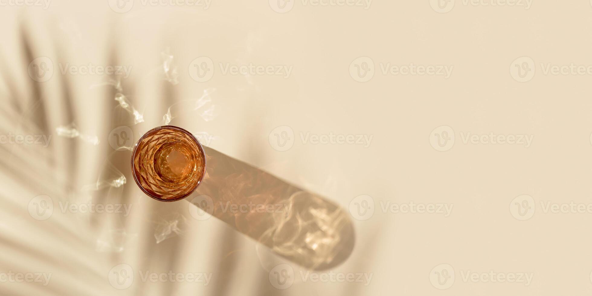 One glass of water from colored glass and beautiful shadows from palm leaf and glare at sunlight, summer drink concept, trend glass orange color, beige background, aesthetic top view photo