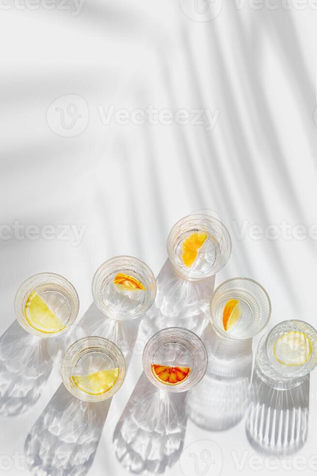 Fruit Infused Water in transparent Glasses, palm leaf Shadow at sunlight on White background with caustics effect. Aesthetic summer fresh drink with slices of lemon and orange in glass cups, top view photo