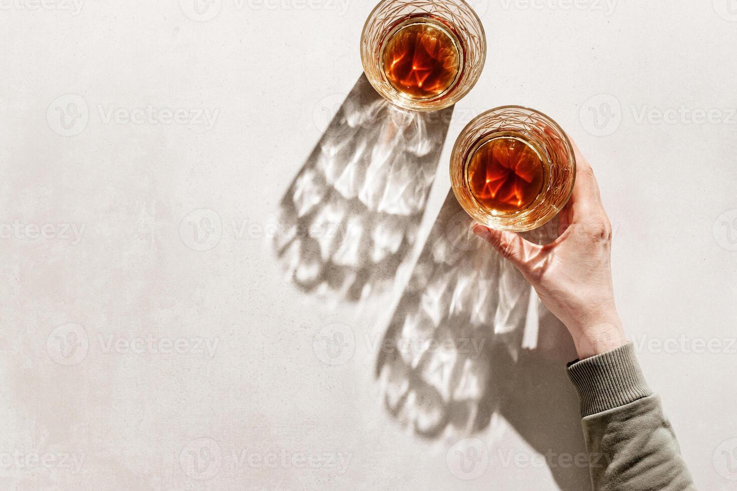 Two Whiskey Glasses with Shadows and sunlight Reflection on beige background, hand holding glass with alcohol, aesthetic top view summer still life, refraction at crystal shine cups, caustics photo