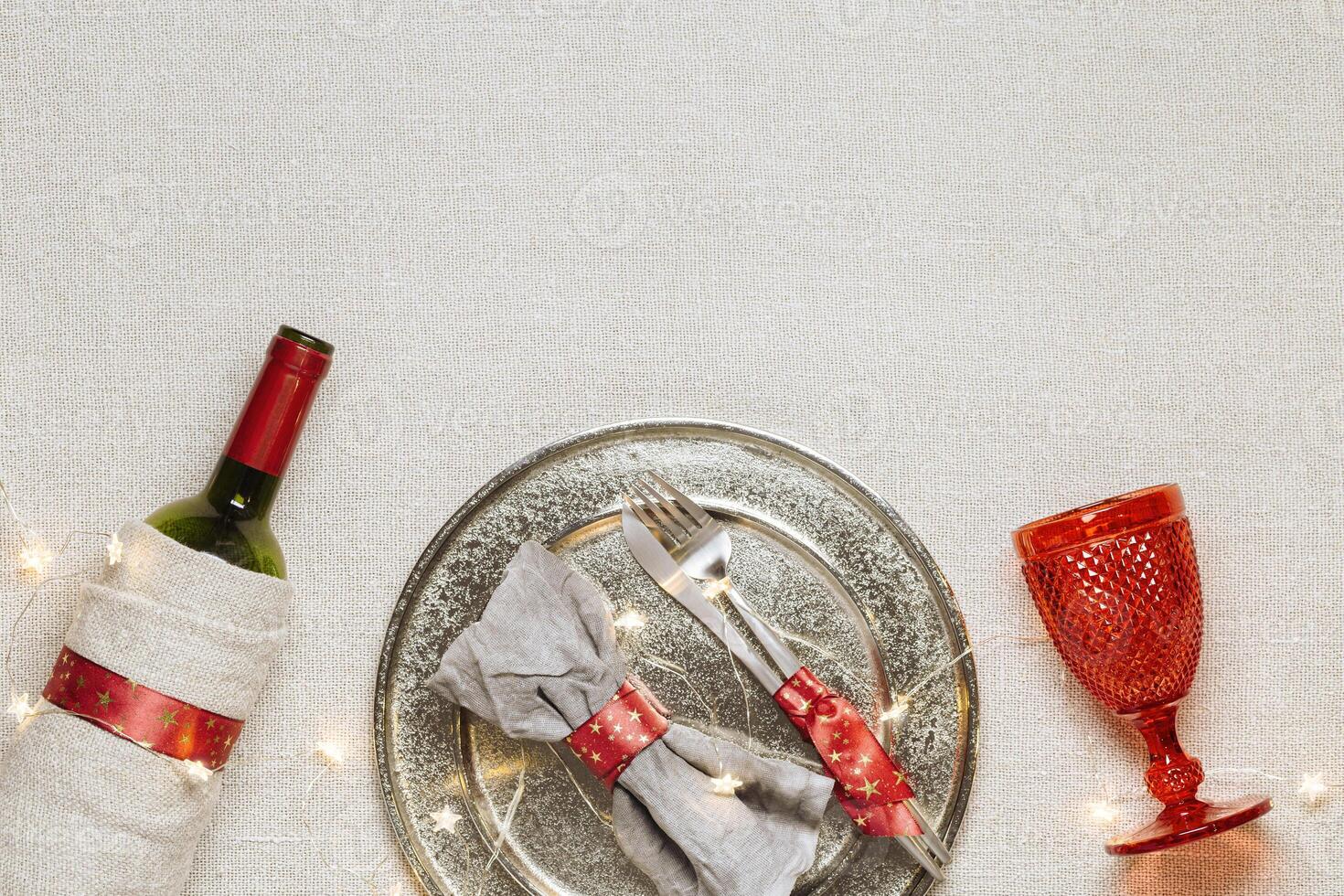 Christmas rustic table setting, red wine glass, plate, knife, fork, wine bottle, decorated green fir tree on sacking tablecloth. New Year dinner with xmas decor, serving festive table photo