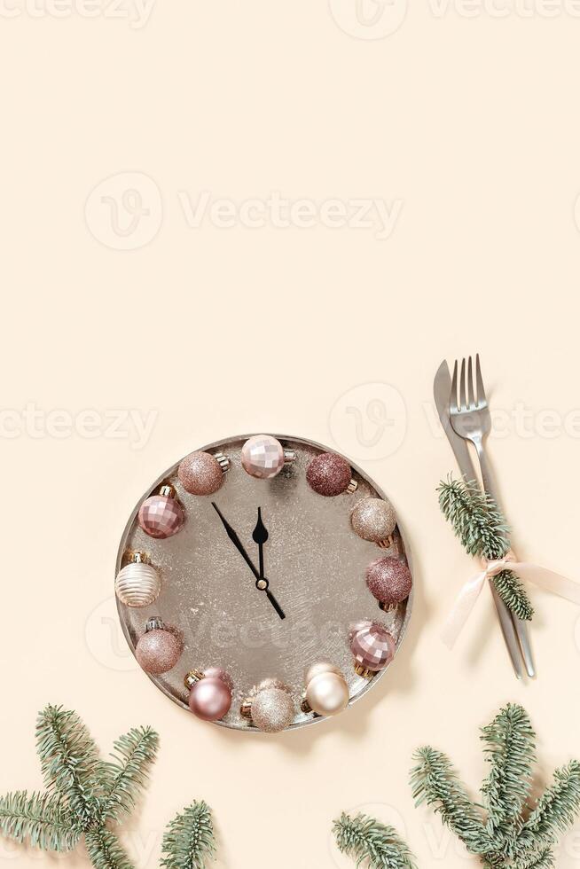 Winter festive table setting concept, Christmas plate with clock hands show twelve or midnight, sparkle balls beige pink color, knife, fork, fir tree branches. New Year holiday food concept photo