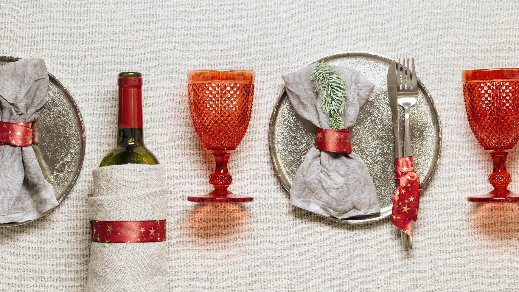 Tableware for Christmas or Xmas festive dinner, linen napkin on plate, knife, fork, red wine glass, bottle of wine on rustic tablecloth, New Year holiday cutlery, red decor, overview photo