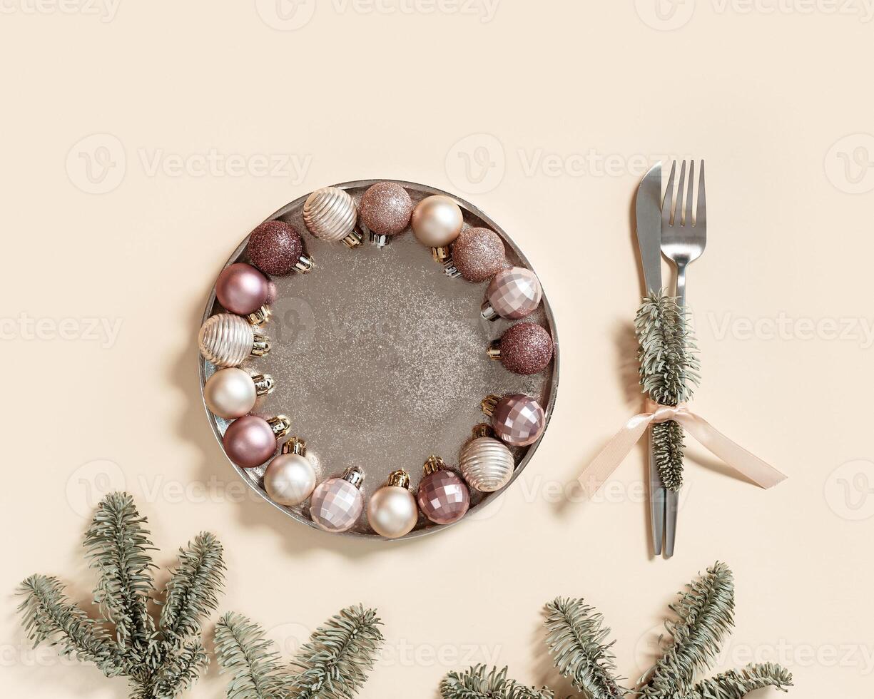 Christmas table setting concept, shining small Christmas balls beige pink color in metal plate, knife, fork decorated green branches fir tree, New Year holiday dinner or party idea photo