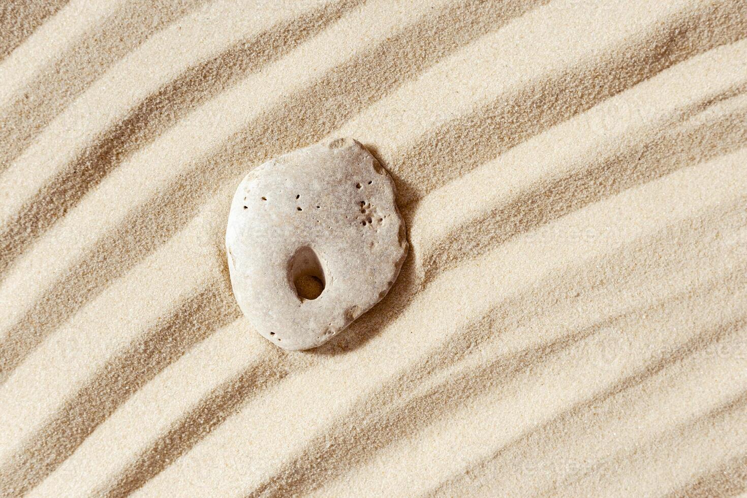 Minimal background with one stone with hole on fine sand with wave ripple. Natural happy stone light ivory color. Symbol of fun, fortunate, lucky. photo
