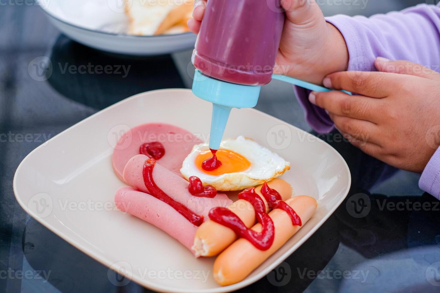 Child hand squeezing ketchup onto a breakfast plate with ham, sausages, and a fried egg A bowl with sliced bread is blurred in the bac photo