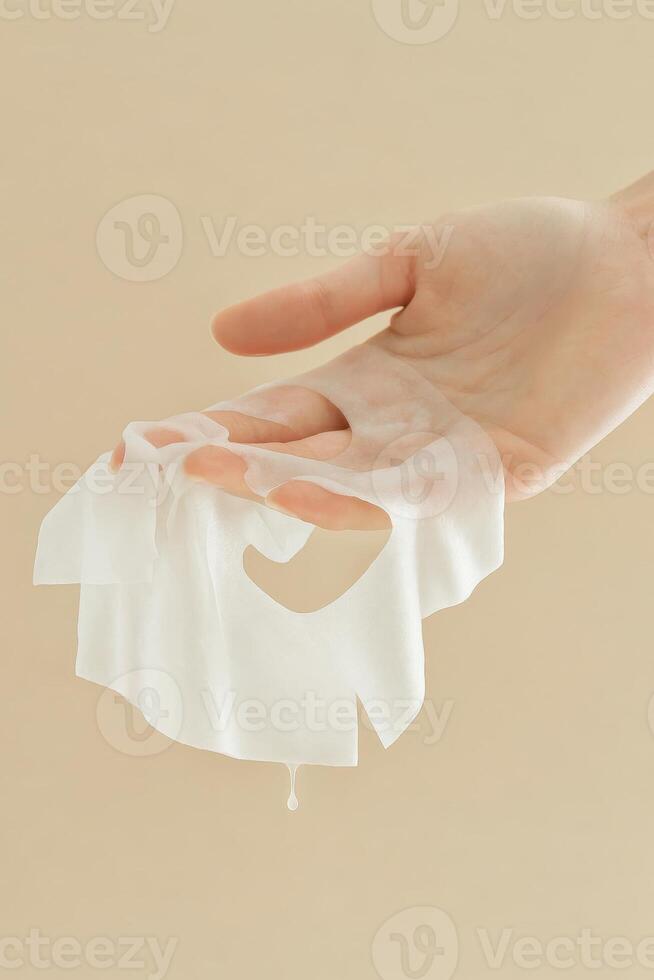 Close-up of a pale hand gently holding a soaked white facial sheet mask, with a visible droplet dripping from the mask. photo