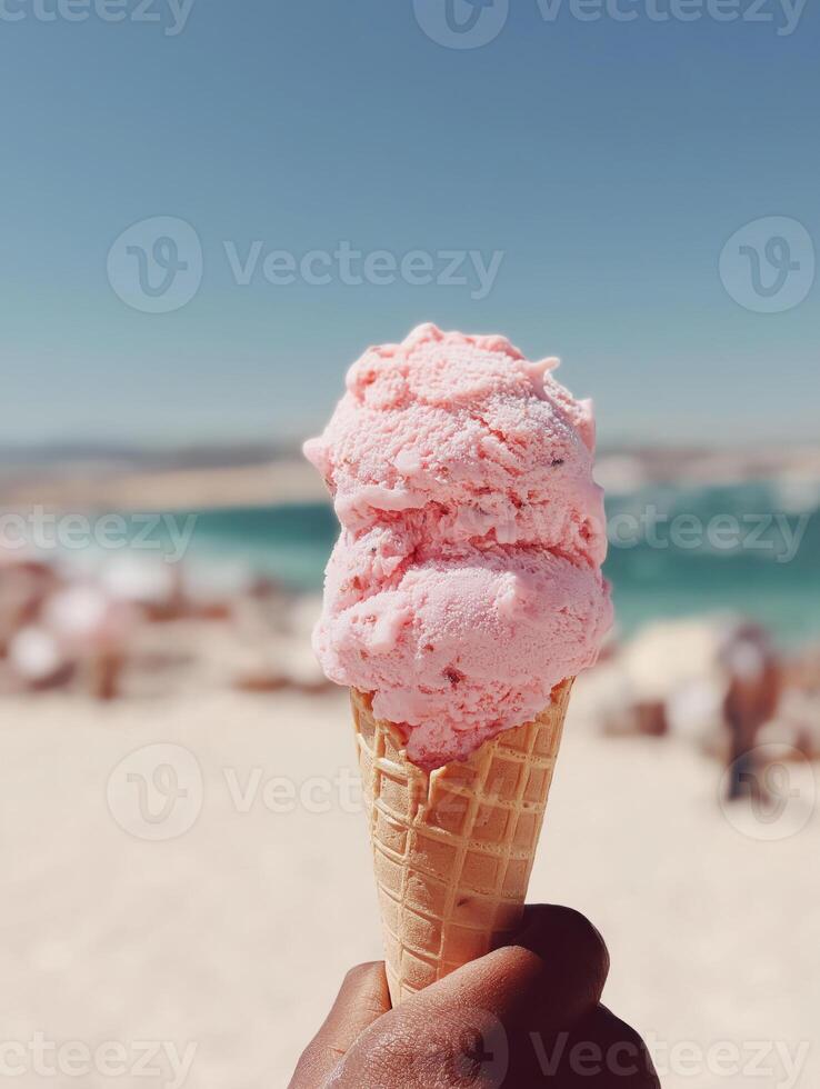 A close-up of a hand holding a pink strawberry ice cream cone against a bright, sunlit beach with blue sea and pale sky in the background. photo