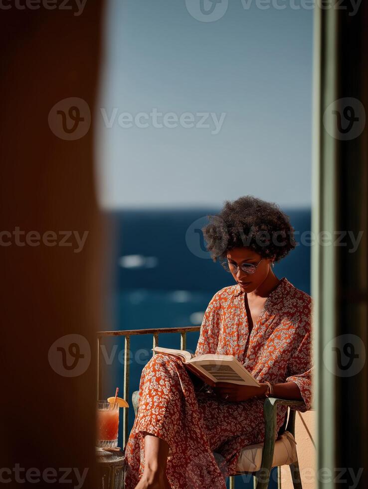 A woman sits in a wicker chair at a round table on a terrace by the sea, holding a book, with a cocktail glass next to her, against the backdrop of a sparkling ocean surface. photo