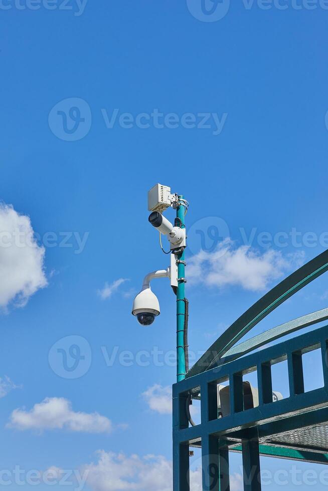 Urban security cameras and monitoring devices installed on a green pole above a public structure, set against a bright blue sky photo