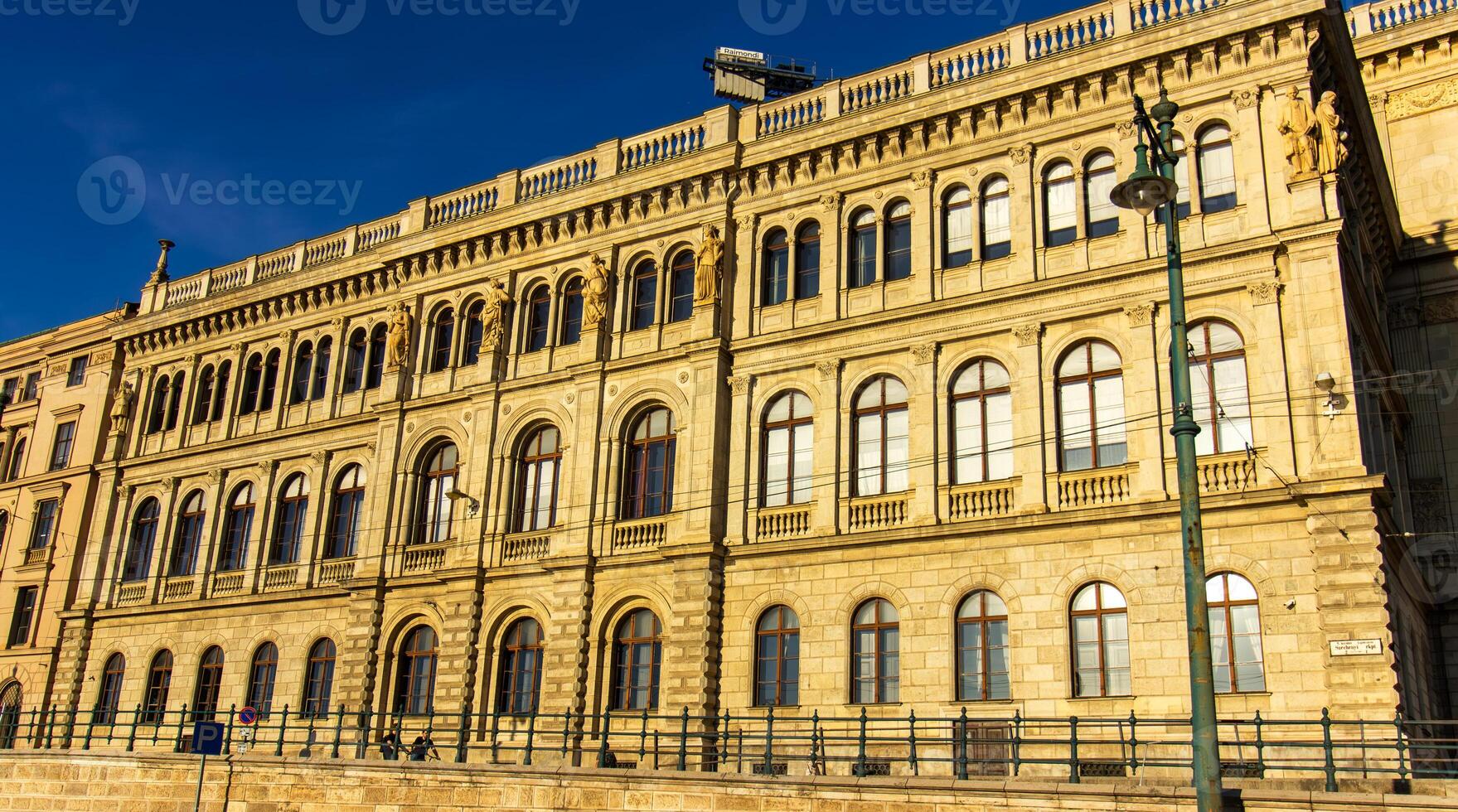 Facade of historic neoclassical building with arched windows and sculptures in Budapest, Hungary, illuminated by warm sunlight under clear blue sky photo