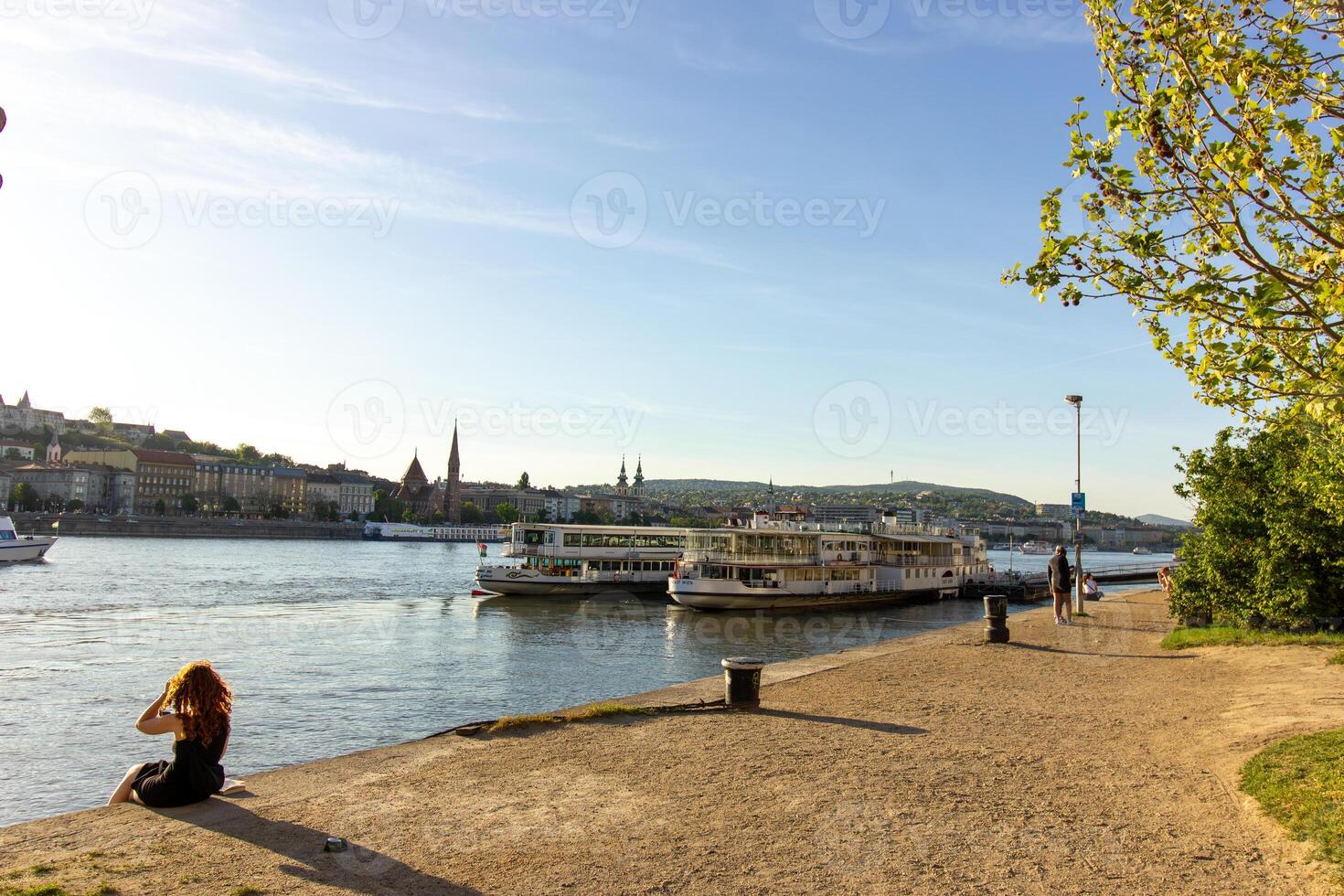 Scenic riverside view of Danube in Budapest with docked cruise boats, historic cityscape in background, and people relaxing on the promenade photo