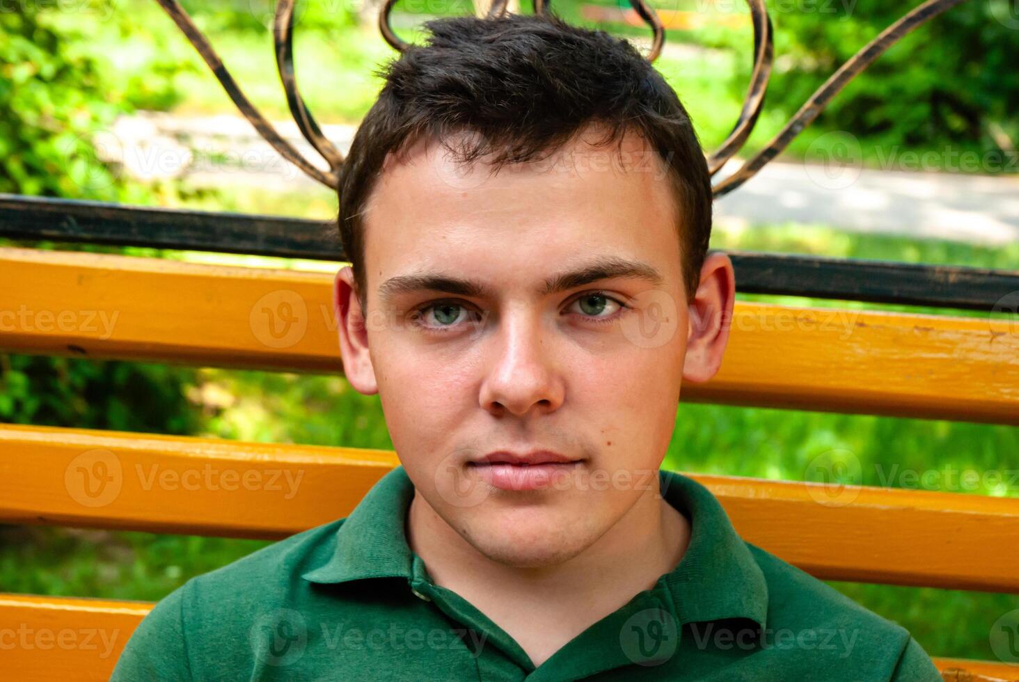 Young caucasian male teenager with brown hair sits on a yellow bench in a lush green park, wearing a green polo shirt, gazing forward intently photo