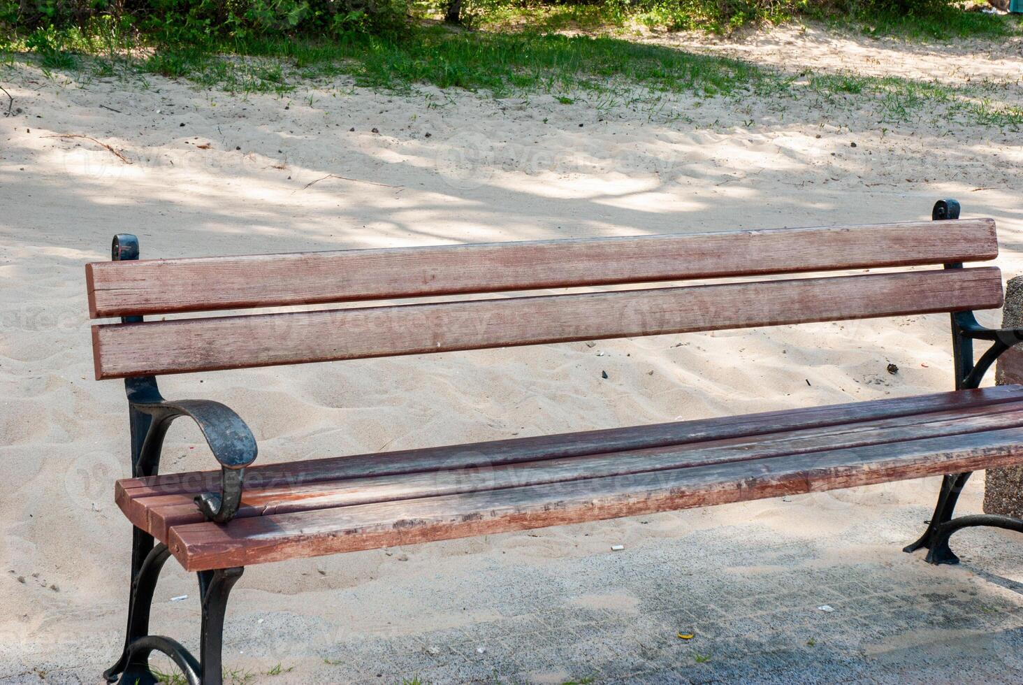 Weathered wooden bench on sandy pathway with elegant iron detailing in sunlit park surrounded by lush greenery and shadows creating serenity. Digital nomad lifestyle. Remote work photo