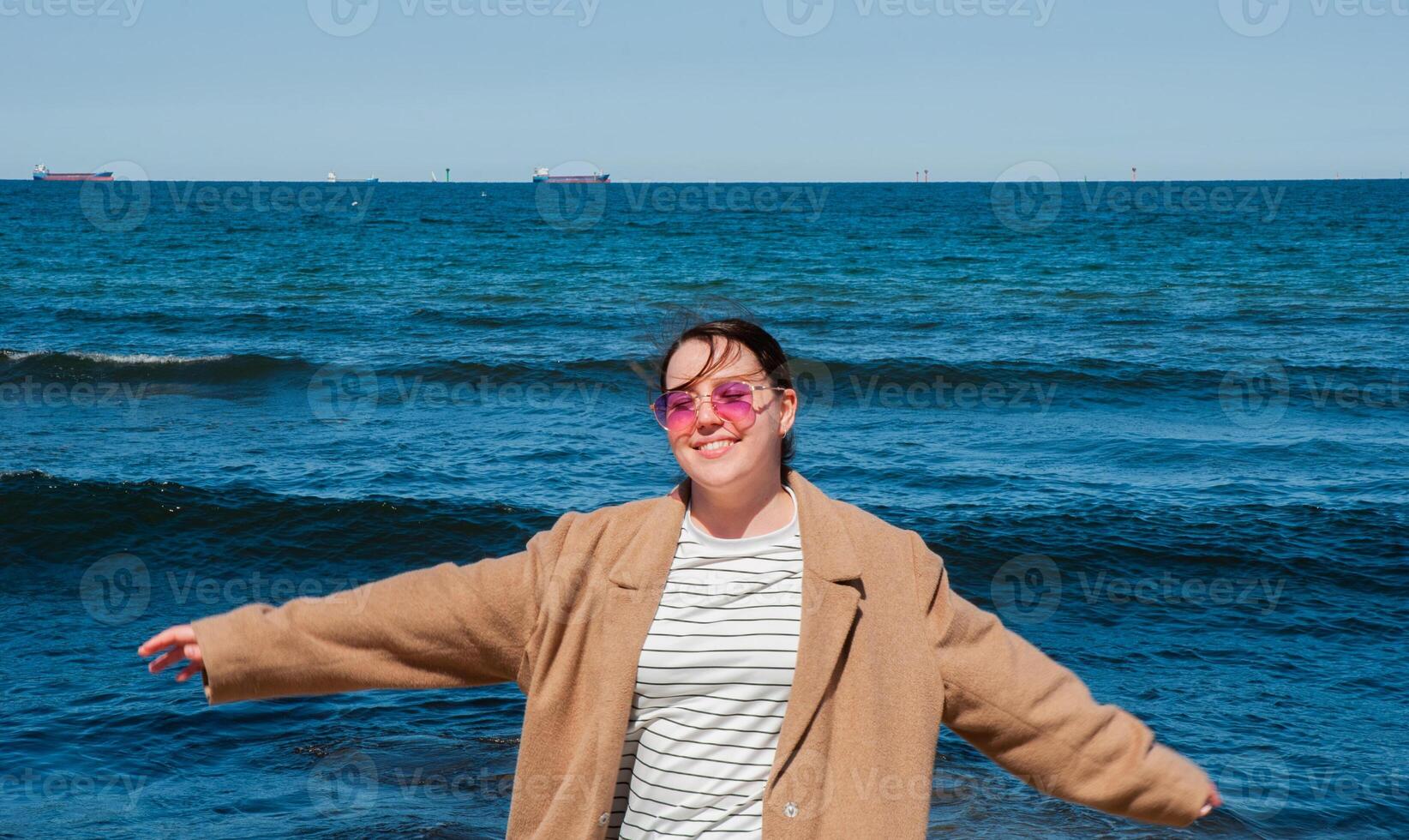 Young caucasian female enjoying a sunny day at the beach, wearing a brown coat, with arms outstretched and pink sunglasses, ocean waves in the background. Digital nomad lifestyle. Remote work photo