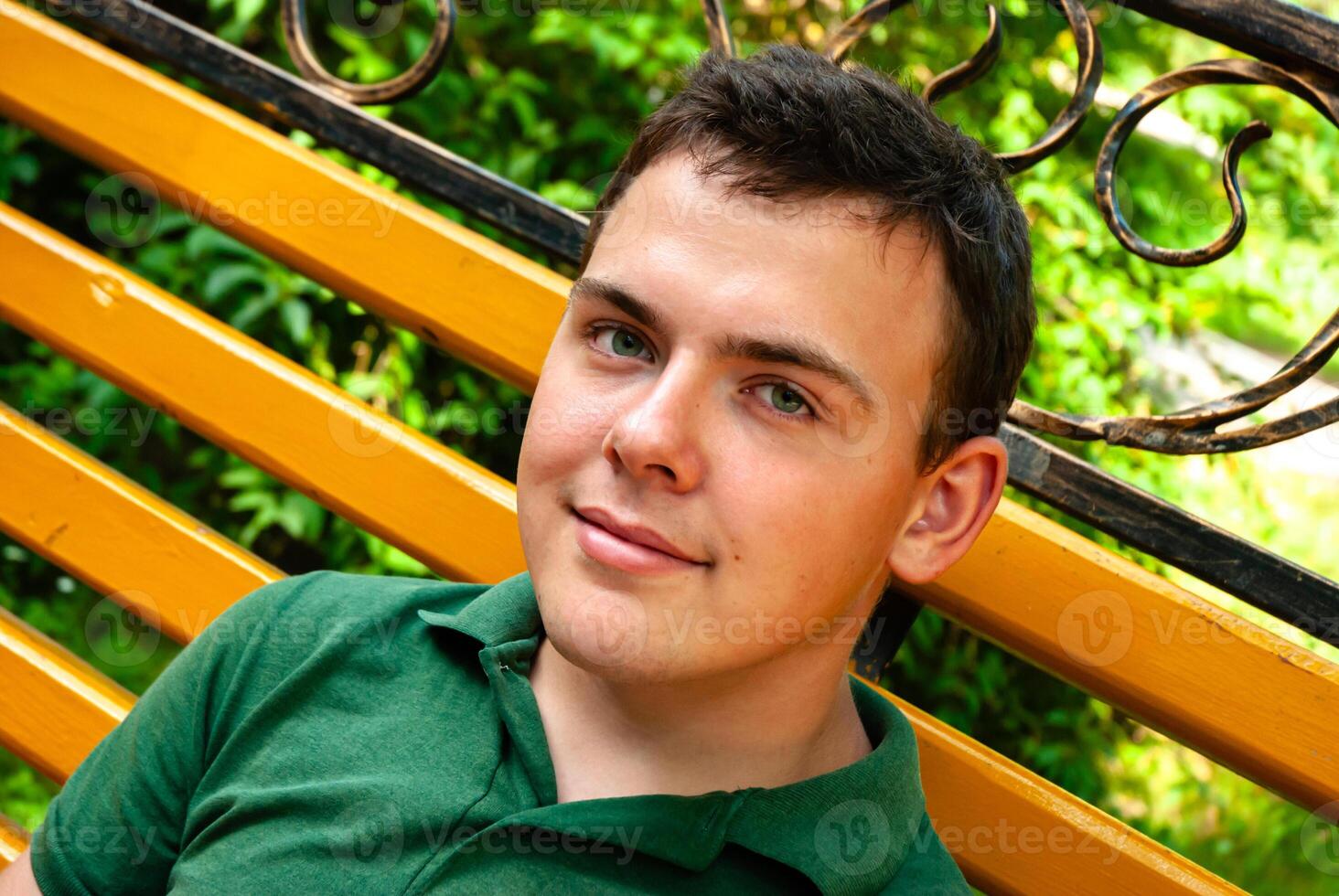 Young caucasian male relaxing on bright yellow bench in a lush green park setting, wearing a green shirt and smiling with contentment photo
