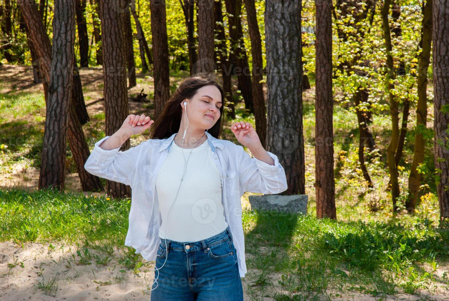 Young caucasian woman enjoying music outdoors in a serene forest setting, wearing casual attire with a relaxed expression and earphones. Digital nomad lifestyle. Remote work photo