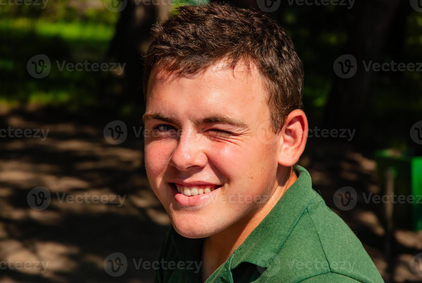 Young caucasian male winking outdoors in green shirt surrounded by natural sunlit background with trees and shadows capturing a happy moment photo