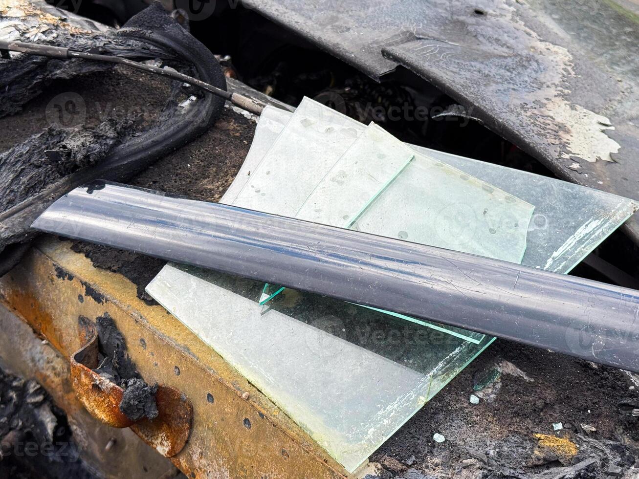 Broken window glass from a car resting on a burned and rusted door frame, with visible soot, melted rubber seals, and fire-damaged metal components photo
