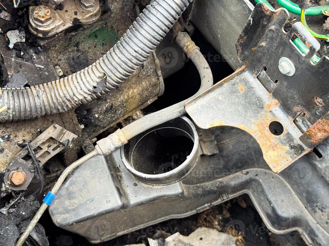 Car radiator neck and mounting bracket showing exposed coolant inlet, rusted metal support, and surrounding engine bay components after fire and mechanical damage photo