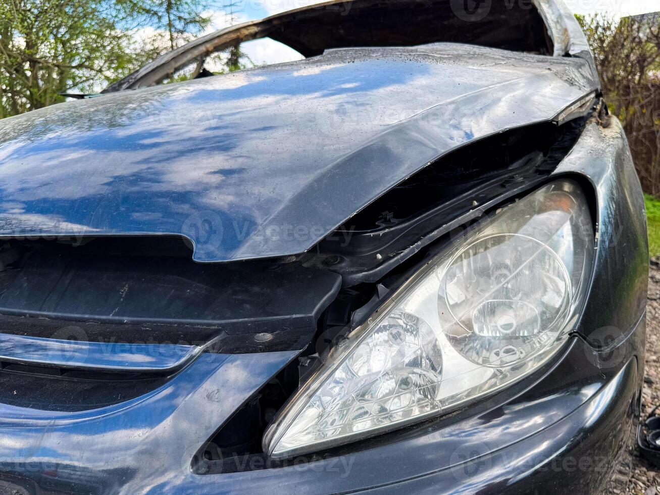 Close-up of a damaged black car front with a broken hood and intact headlight, reflecting the sky and trees in a rural outdoor setting after a fire photo