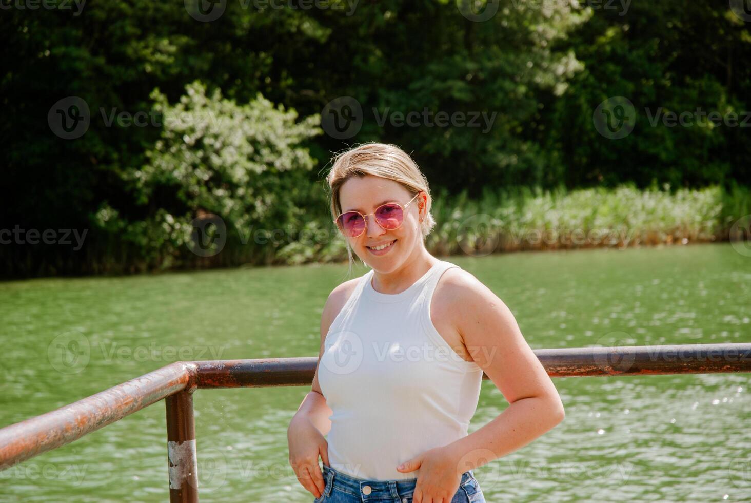 Smiling woman in sunglasses by a tranquil lake in summer. Weekend concept near the forest and lake photo