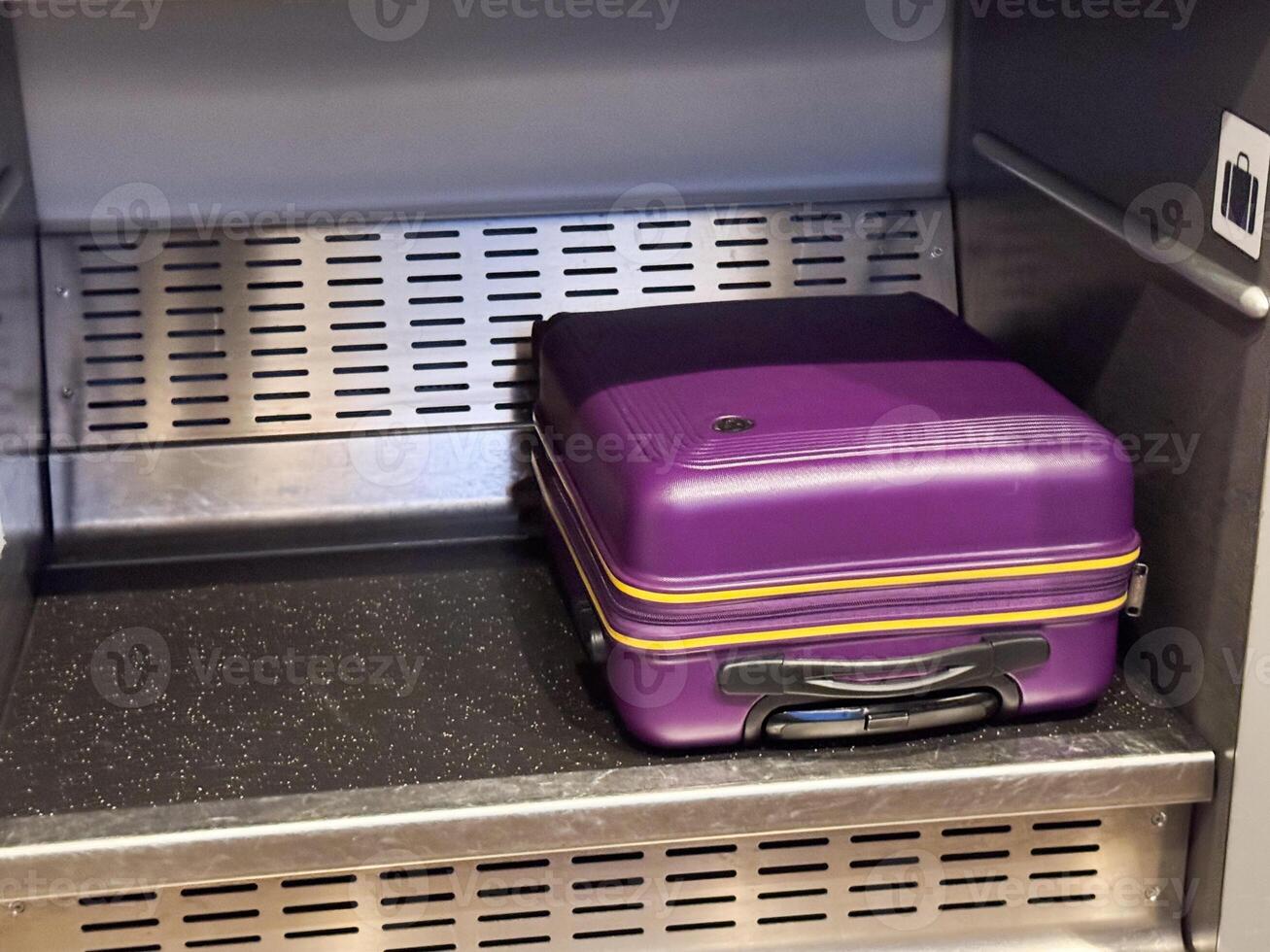 A close-up of a vibrant purple hard-shell suitcase with yellow accent stripes, placed on a lower metallic shelf inside a modern train's luggage compartment photo