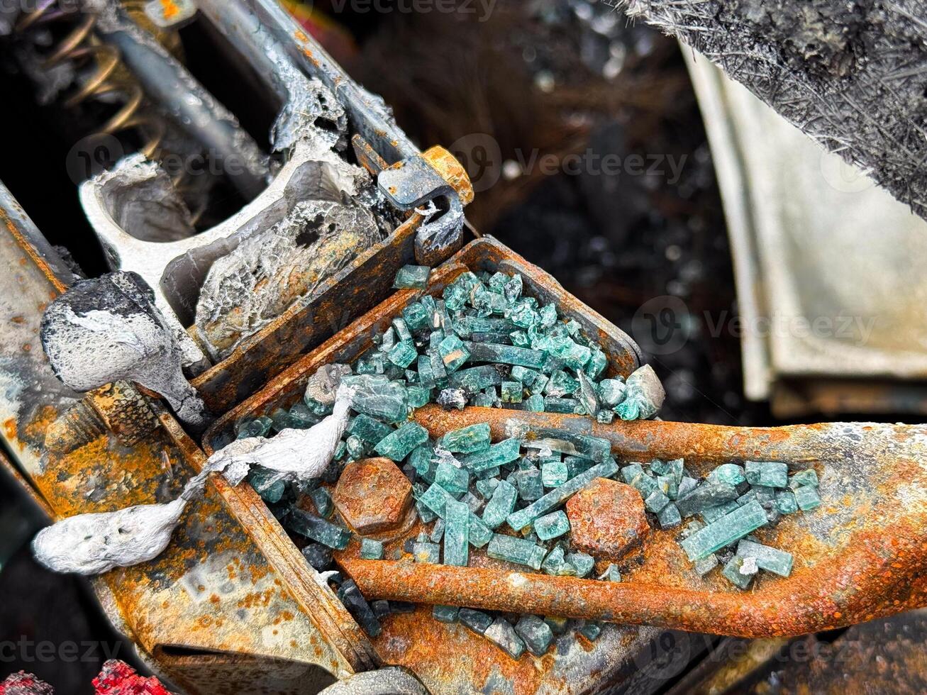 Close-up of shattered automotive safety glass scattered over a burned and rusted metal car seat frame, with visible melted elements and fire damage photo