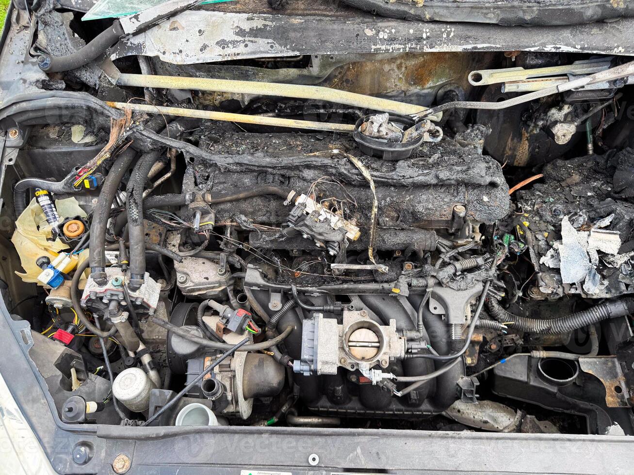 Burned car engine compartment showing fire-damaged engine block, melted wiring, scorched hoses, and exposed intake manifold with severe thermal destruction photo