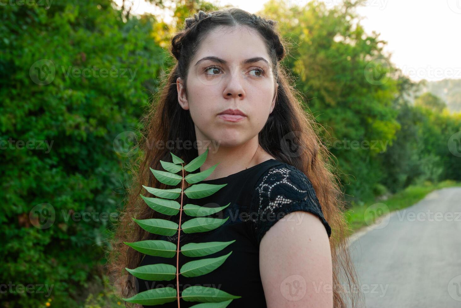 Young caucasian female with long brown hair holding a green leaf on a serene country road surrounded by lush green trees in late afternoon light photo