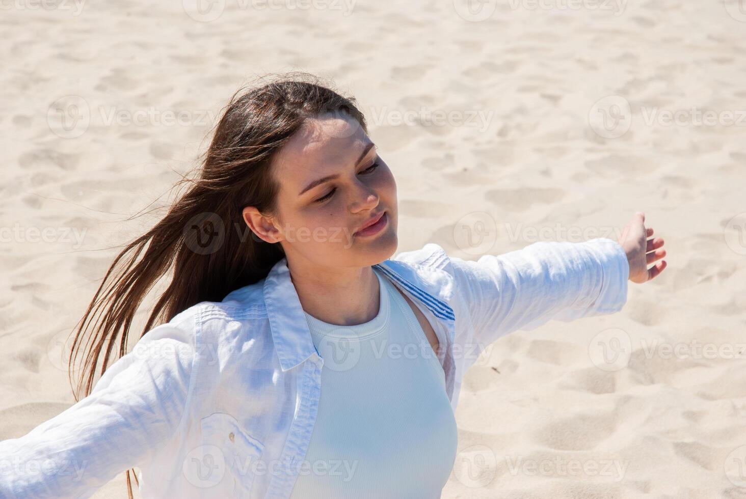 Young caucasian teen girl embracing sunlight on sandy beach with arms outstretched, enjoying a sunny day with relaxed and carefree expression. Digital nomad lifestyle. Remote work photo