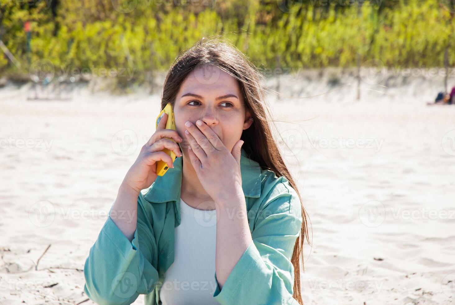 Young caucasian woman at the beach in a green jacket covers her mouth while talking on a yellow phone, expressing surprise and contemplation. Digital nomad lifestyle. Remote work photo