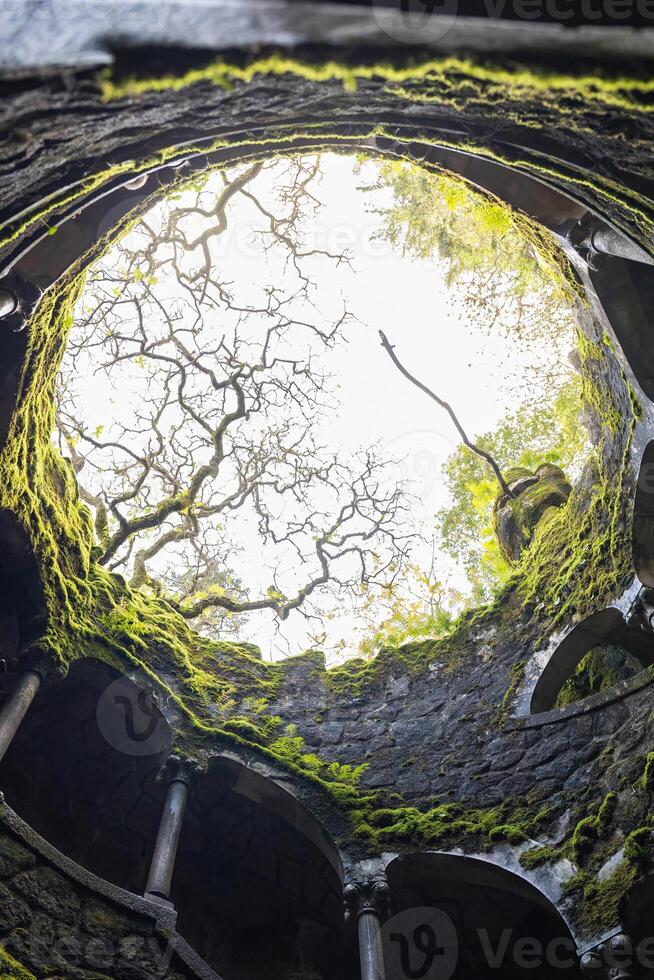 Sintra, Portugal - 02,12,2025 - Visitors explore the Poco Iniciatico of Quinta da Regaleira, a spiral stone well with moss-covered arches and ancient masonry. photo