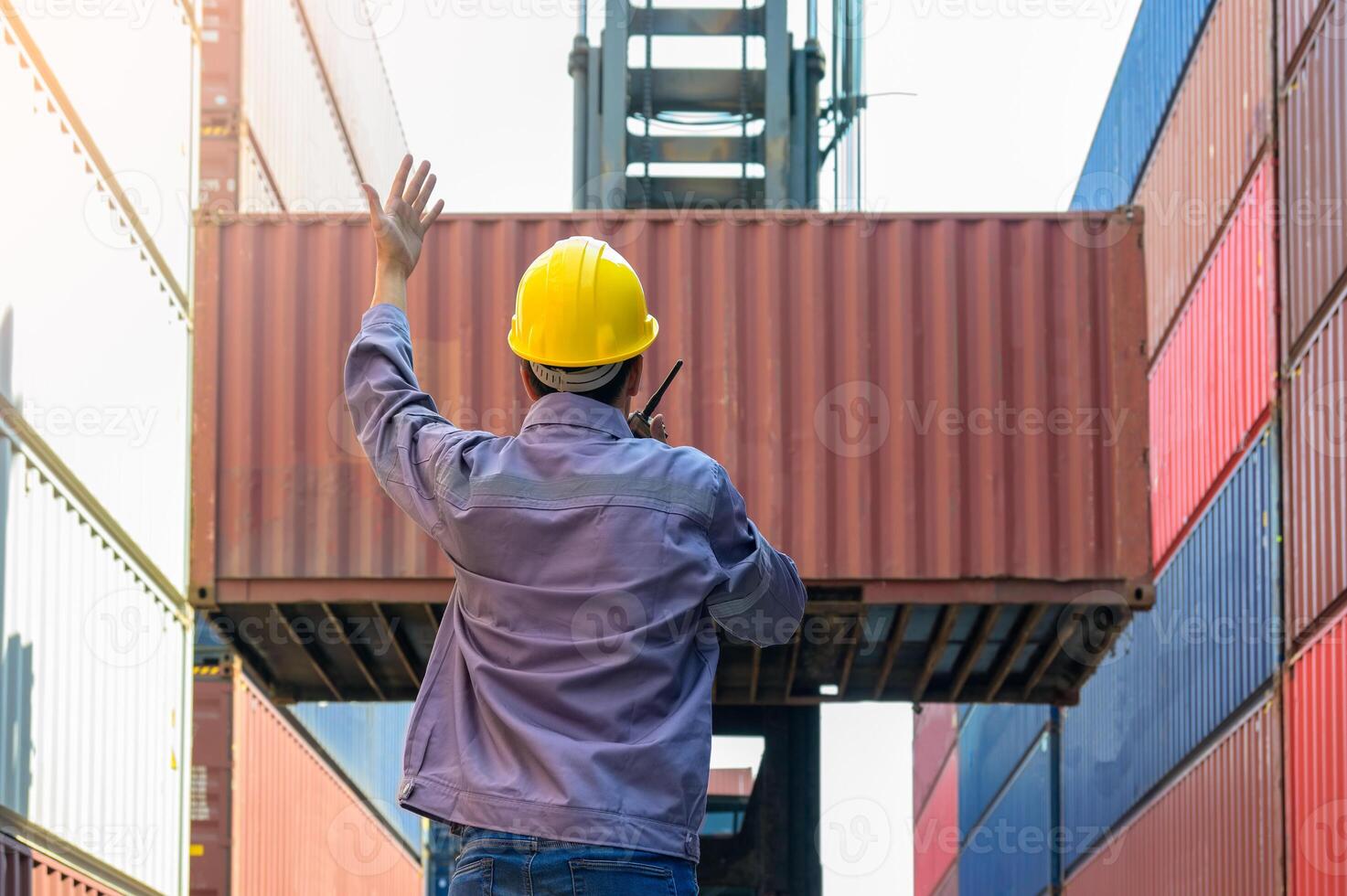Dock supervisor in yellow hard hat and uniform directing container loading with walkie talkie and hand gesture at shipping container yard. photo