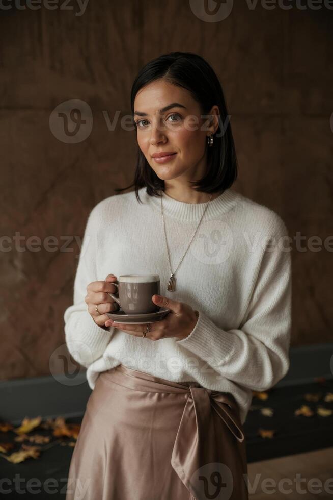 Cozy indoor moment with a woman enjoying coffee while dressed elegantly in a warm sweater and satin skirt during autumn photo