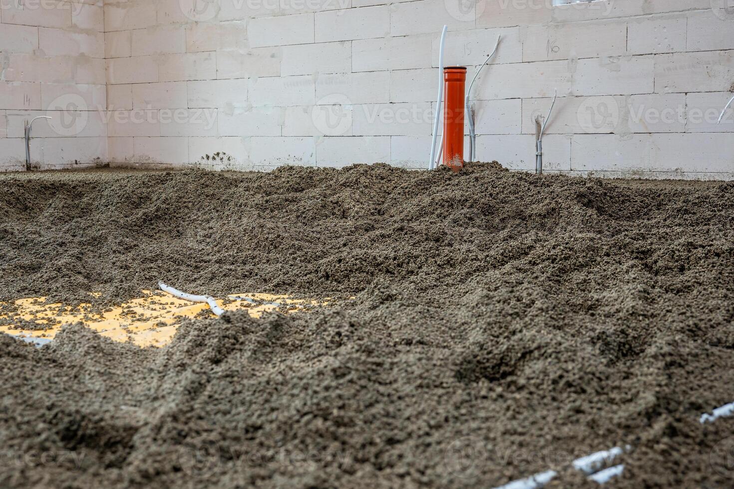 Interior of a construction site featuring an orange vertical pipe, white conduit pipes, loose gravel floor, and light colored concrete block walls. photo