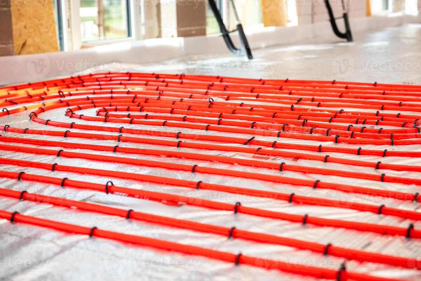 Bright red underfloor heating pipes arranged in loops over reflective insulation, secured with black fasteners, in a well lit construction site interior. photo