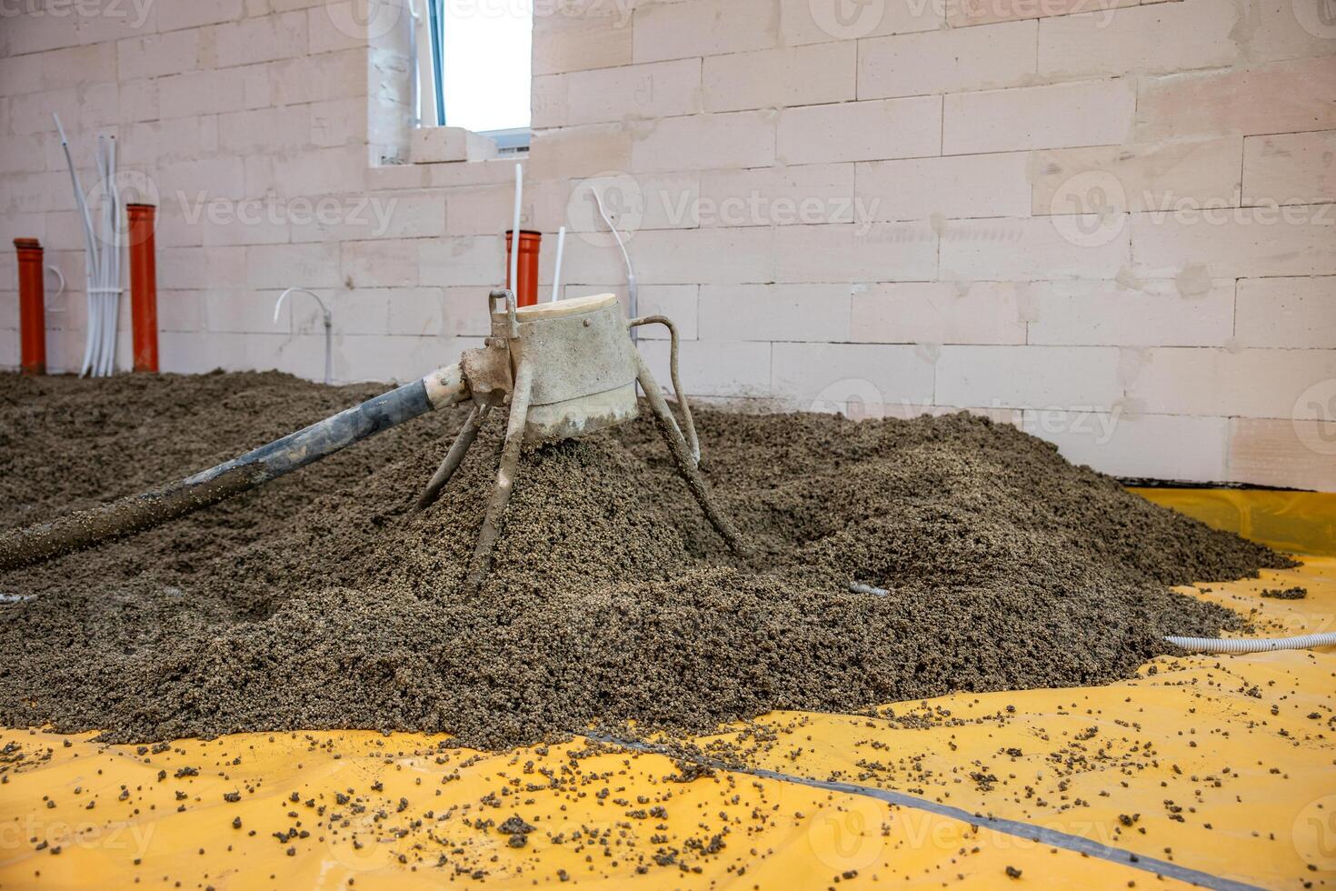 A construction site showing lightweight aggregate spread on a yellow vapor barrier. A mechanical tool disperses material near concrete walls and utility pipes. photo