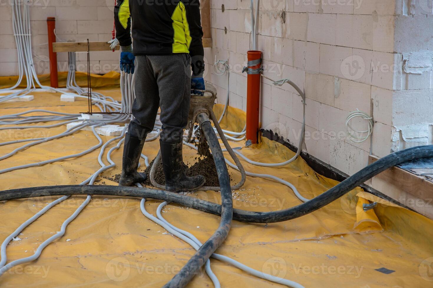 A worker operates equipment to pour concrete over underfloor heating pipes on a yellow sheet in an indoor construction site with unfinished walls. photo