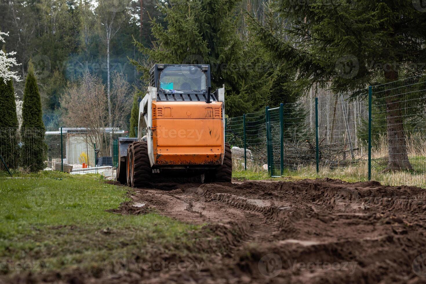 An orange and white skid steer loader works on a dirt path, leaving tire tracks. A green metal fence, tall evergreen trees, and a structure with a transparent roof are visible. photo