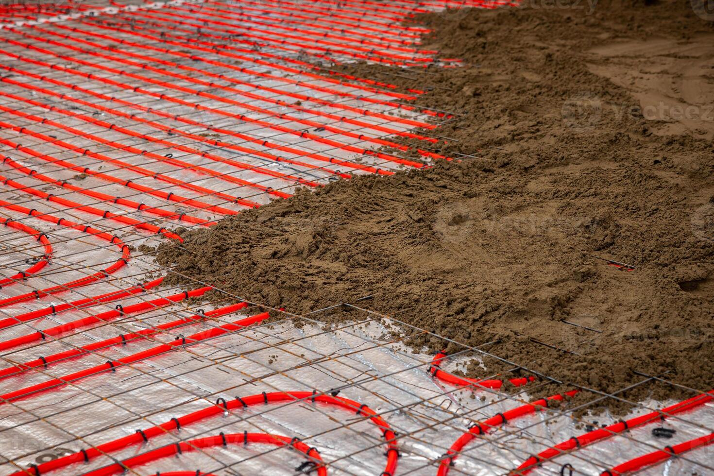 Red underfloor heating pipes are installed over reflective insulation, secured by a metal grid, with Estrich concrete partially covering the pipes. photo