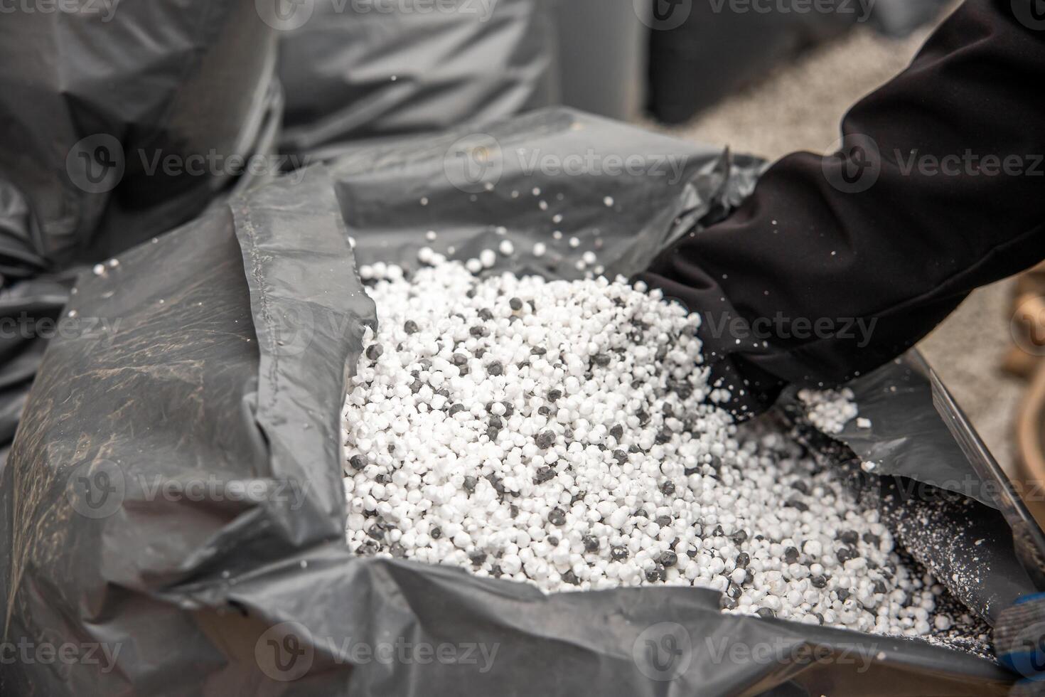 A gloved hand reaches into a black plastic bag containing white and gray granular material, with additional bags and a gravel surface in view. photo