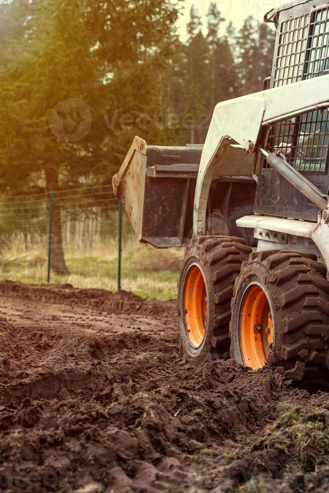 A compact loader with orange wheels and raised bucket on muddy ground. A wire fence and tall evergreen trees are visible in the outdoor setting. photo