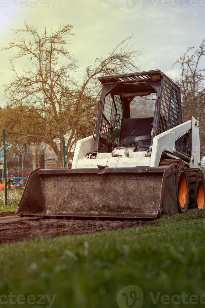 A white skid steer loader with orange wheels sits on a dirt patch surrounded by grass. Leafless trees and a faint fence are visible in the background. photo