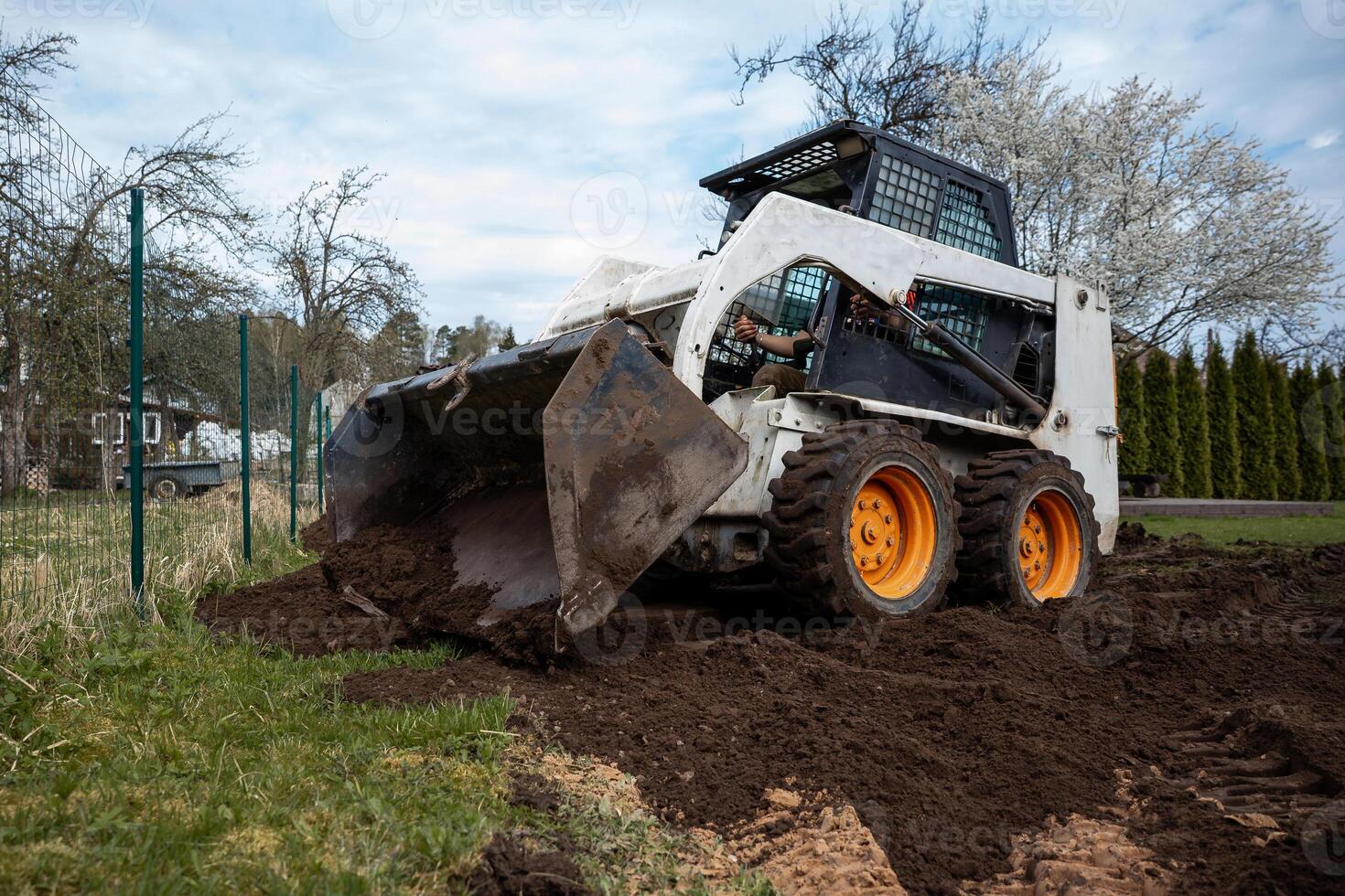 A skid steer loader with orange wheels moves soil near a wire fence. Trees, some in bloom, and tall shrubs surround the outdoor work area. photo